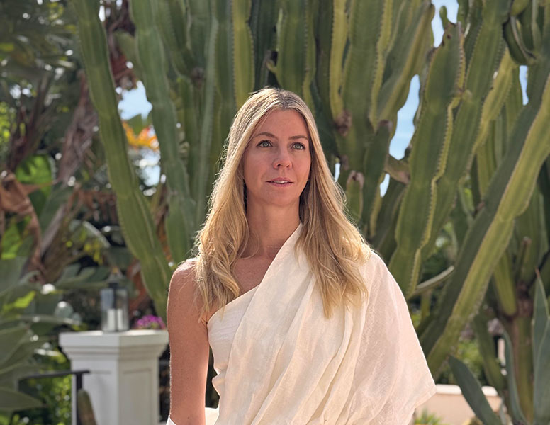 Woman with long blonde hair wearing a white sari standing outdoors in front of tall cactus plants.