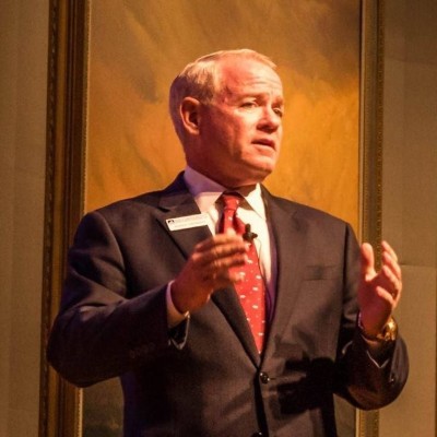 Older man in a dark suit and red tie giving a presentation, gesturing with his hands on a stage.